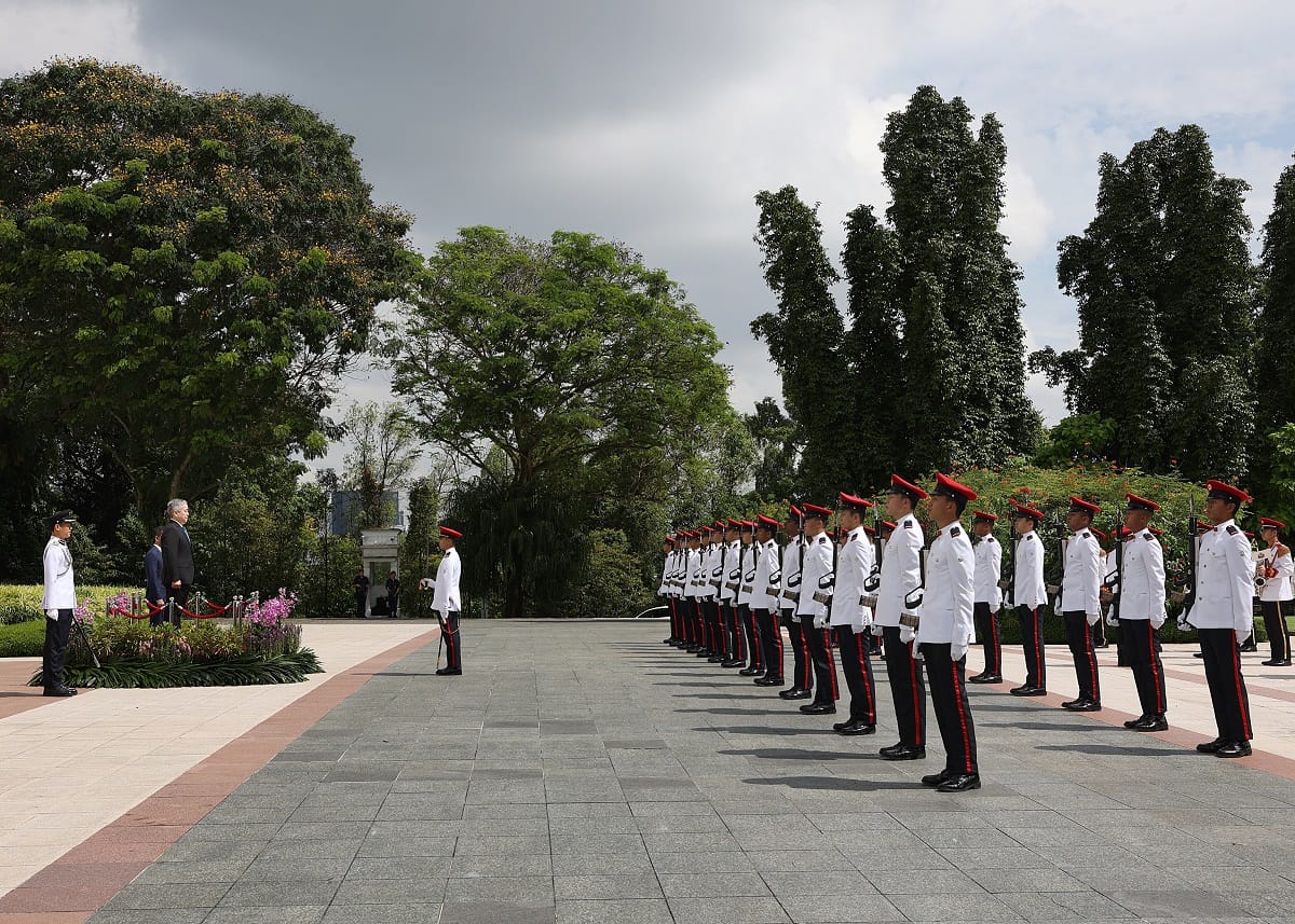 Honor guard standing in formation in white uniforms with red trim, facing a dignitary.